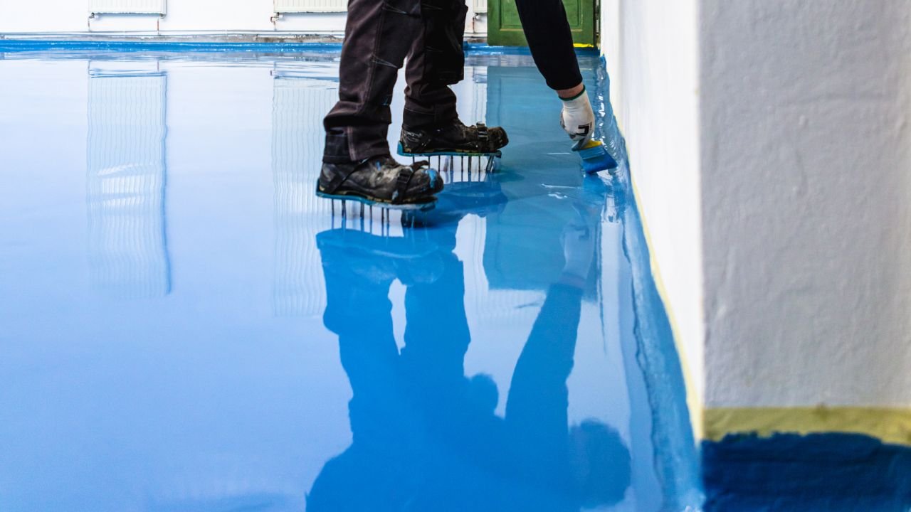 Applicator in spiked shoes brushing blue epoxy coating along a wall edge