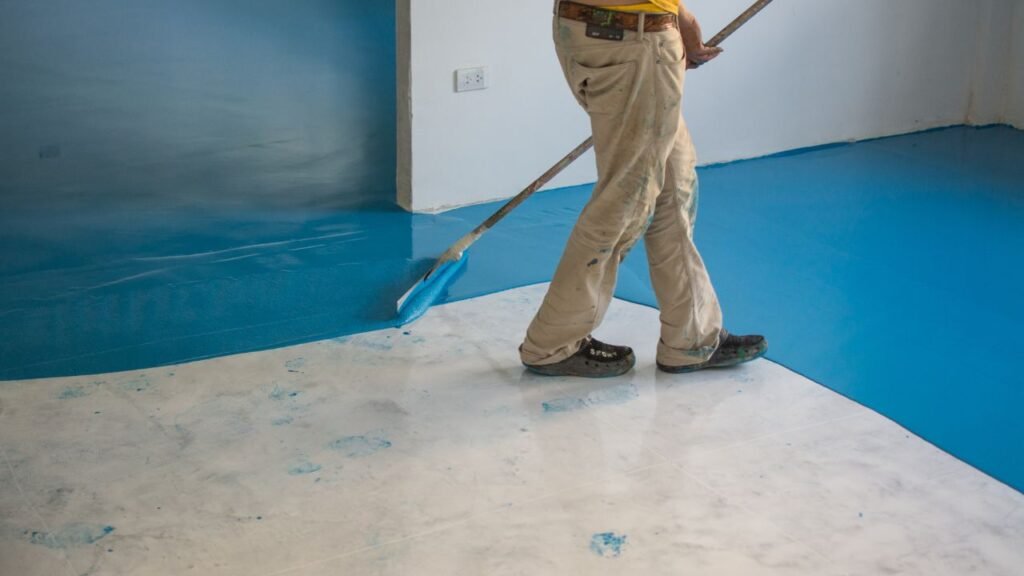 Contractor spreading blue epoxy floor coating with a squeegee in empty room