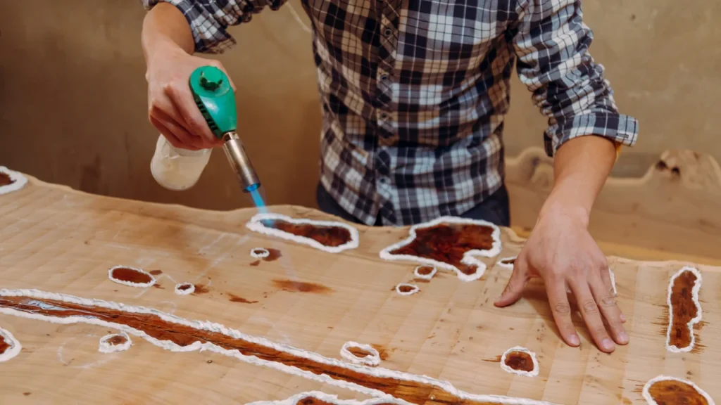Craftsman using a heat torch to remove air bubbles from epoxy resin filling cracks in a live-edge wood slab
