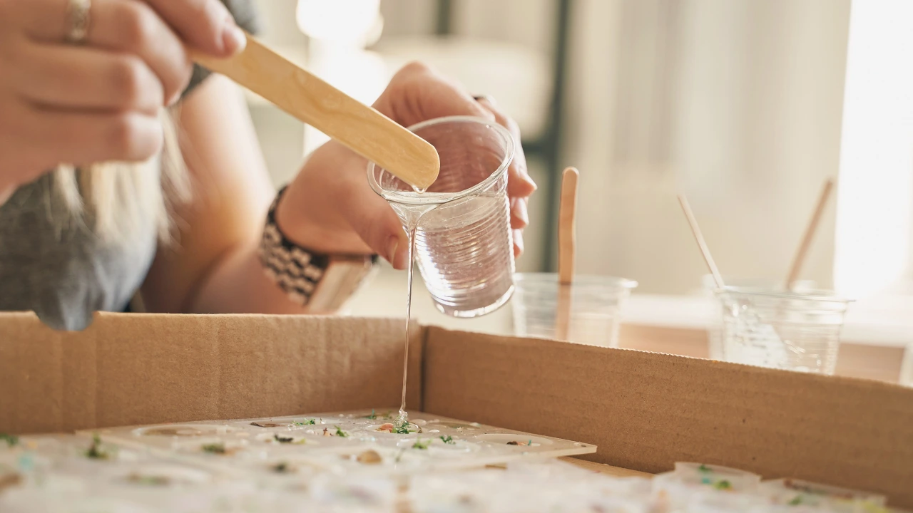 Person mixing clear casting resin in a plastic cup with a wooden stir stick over a craft mold