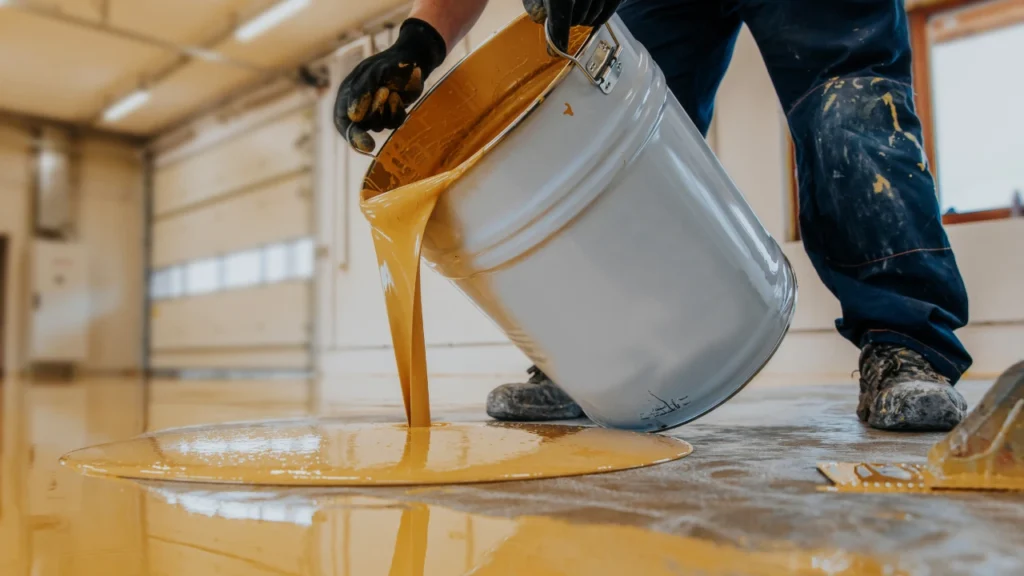 Worker in gloves pouring liquid epoxy resin from a bucket onto a garage floor for coating