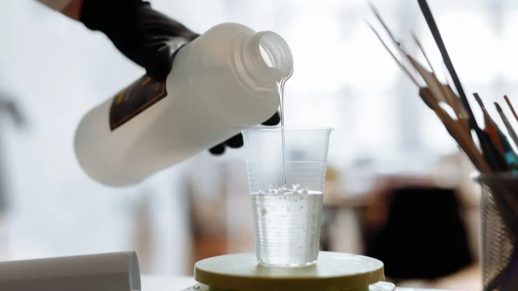 Gloved hand pouring isopropyl alcohol into a plastic cup, used to dissolve uncured resin