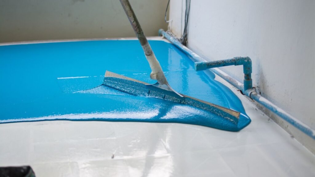 Notched squeegee spreading teal epoxy floor coating near wall pipes